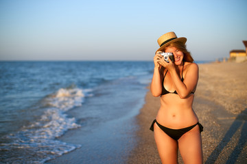 Young authentic woman with vintage retro film camera enjoying tropical beach on summer vacation. Female travel photographer in straw hat taking photos having fun at sea. Real girl unretouched shape.