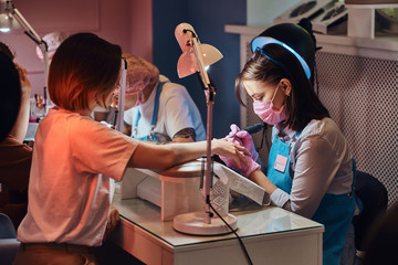 Young attractive manicure master is doing nail treatment for another woman at busy nail studio.