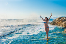 A Girl In A Swimsuit, Splashing From The Waves, Brunette Girl Rejoices Splashing From The Waves. Young Happy Woman Jumping With Raised Arms, Waving A Blue Bikini Swimsuit In A Blue Swimsuit And A