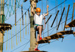 © Natali - Boy in forest adventure park. Kid in orange helmet and white t shirt climbs on high rope trail. Climbing outdoor, amusement center for children. Young boy plays outdoors