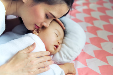 Wall Mural - Young pretty Asian mother hugging her sleeping cute baby on the bed. The mother closing her eyes when touching her child softly with smile and feeling happy. Love of mother and daughter concept.