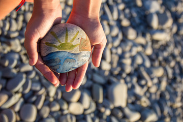 Bright sun painted on pebble in the hands of a child on the background of a pebble beach. Pebbles and sea background.