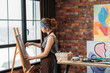 © golubovy - Home art studio. Side view of young female painter drawing on easel. Window, brick wall with abstract artworks in background.
