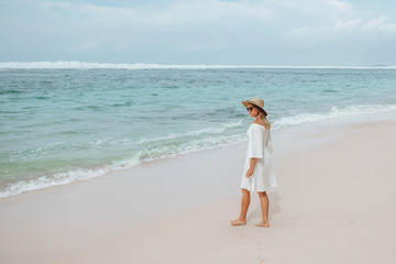 girl in a white dress walks on a white beach in a hat and sunglasses