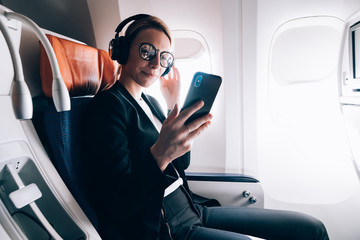 Wall Mural - Travel tourism technology and air flights concept, intelligent woman sitting in plane with modern smartphone gadget and searching favourite music playlist in application for melomans for listening