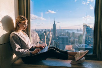 Concentrated female freelancer keyboarding information on laptop computer working remote in coworking office with panoramic views of scenic Manhattan downtown. Student exchange and travel to New York