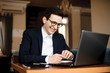 © Strelciuc - Portrait of handsome caucasian manager sitting at his desk laughing while looking at his laptop wearing eyeglasses dressing suit.