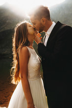 Side View Portrait Of A Amazing Wedding Couple Kissing Against Sunset While Groom Is Touching Bride's Face With A Hand.