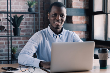 Wall Mural - Portrait of positive Afrcan business man sitting in front of laptop in loft office against red brick wall