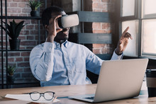 Young african businessman wearing virtual reality headset, pointing in the air with finger while sitting at his desk in modern office