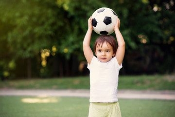 Little soccer player standing at football field outdoors: toddler boy holding ball above head ready to throw it in sunny day. Summer outdoors fun and sports games for kids concept. Copy space