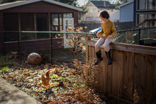 A happy child in yellow sits on a backyard fence smiling at a chicken