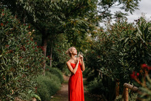 Side View Of Attractive Blonde Dressed In Red Sensually Posing With Closed Eyes Among Green Blooming Trees
