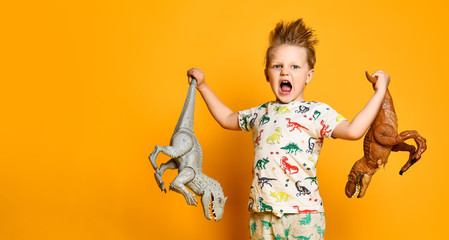 little cheerful boy holds a plastic dinosaur in each hand. The boy is dressed in a suit with pictures of dinosaurs.