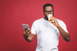 © denis_vermenko - Typing a message. Cheerful black man typing something on the mobile phone, drinking coffee while standing isolated against red background.