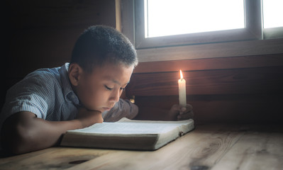 Wall Mural - Christian boy readidng bible with light of candle on wooden table near the window at home. focus at face.