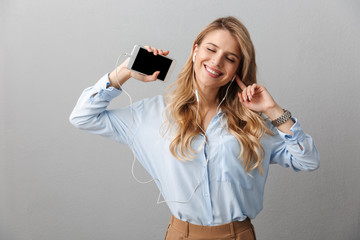 Sticker - Photo of charming blond businesswoman with long curly hair rejoicing while listening to music with smartphone and earphones