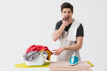 Wall Mural - Young confused displeased houseman househusband with iron near clothes isolated over white wall background talking by mobile phone.