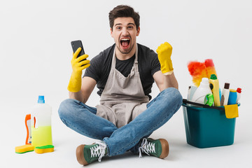 Wall Mural - Excited young houseman househusband with cleansers on floor isolated over white wall background make winner gesture using phone.