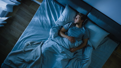 Top View of Handsome Young Man Sleeping Cozily on a Bed in His Bedroom at Night. Blue Nightly Colors with Cold Weak Lamppost Light Shining Through the Window.