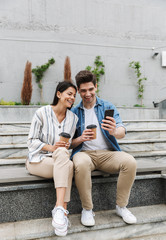 Poster - Image of excited couple drinking takeaway coffee and using smartphone on city stairs outdoors