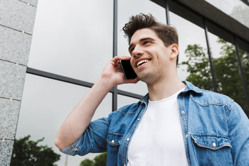 Poster - Happy young amazing man businessman posing outdoors outside walking talking by mobile phone.