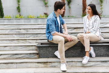 Wall Mural - Image of young couple talking and smiling while sitting on bench near stairs outdoors