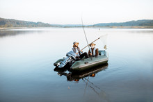 Grandfather With Adult Son Fishing On The Inflatable Boat On The Lake With Calm Water Early In The Morning. Wide Landscape View
