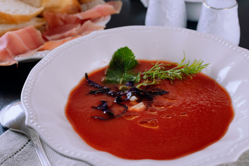 Cold tomato soup gaspacho in a porcelain plate next to sliced ​​jamon, bread and vegetable ingredient on a dark table