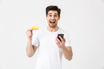 Wall Mural - Excited happy young man posing isolated over white wall background using mobile phone holding credit card.