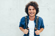 Leinwandbild Motiv Outdoors horizontal portrait of young man with curly hair, smiling broadly, with backpack on the back, standing at building concrete background on the city street. People and lifestyle.
