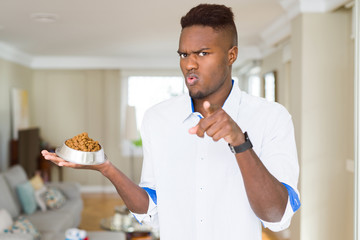 Sticker - African american man holding metal bowl with cat or dog dry food pointing with finger to the camera and to you, hand sign, positive and confident gesture from the front