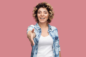 Portrait of happy young woman with curly hairstyle in casual blue shirt standing, toothy smile, looking and giving her hand to greeting and handshake. indoor studio shot, isolated on pink background.