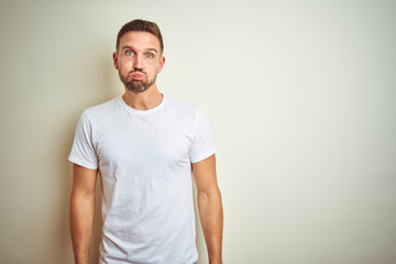 Poster - Young handsome man wearing casual white t-shirt over isolated background puffing cheeks with funny face. Mouth inflated with air, crazy expression.