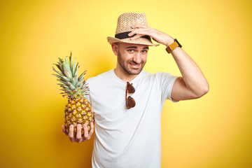 Sticker - Young man wearing summer hat and holding pineapple over yellow background stressed with hand on head, shocked with shame and surprise face, angry and frustrated. Fear and upset for mistake.