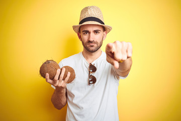 Wall Mural - Young man wearing summer hat and holding coconut fruit over yellow background pointing with finger to the camera and to you, hand sign, positive and confident gesture from the front