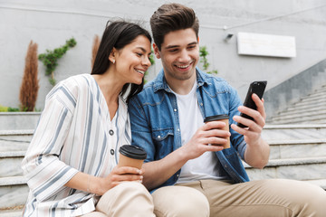 Sticker - Image of cheery couple drinking takeaway coffee and using smartphone on city stairs outdoors