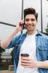 Poster - Happy young amazing man businessman posing outdoors outside walking talking by mobile phone drinking coffee.