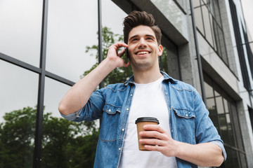 Poster - Happy young amazing man businessman posing outdoors outside walking talking by mobile phone drinking coffee.