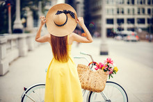A Cute And Stylish Girl In A Yellow Dress And Long Hair Rides On A Bicycle With A Basket Of Flowers In The Sunny Summer City