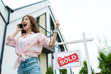 Wall Mural - low angle view of cheerful woman gesturing while talking on smartphone near sold lettering on board