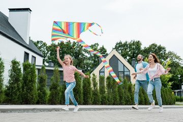 Wall Mural - low angle view of happy kid running with colorful kite near cheerful parents on street