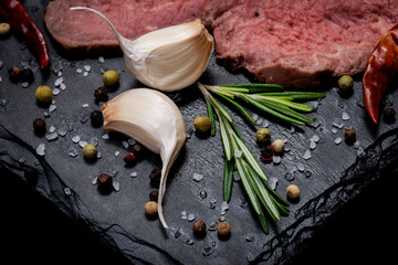 Wall Mural - Grass Fed Corn Roast Beef garnished with Fresh Rosemary, dried Red Chile Pepper, Garlic and Rainbow  Peppercorns on natural black stone background.