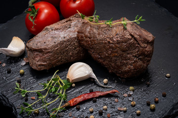 Wall Mural - Grass Fed Corn Roast Beef garnished with Tomatoes, Fresh Thyme, dried Red Chile Pepper, Garlic and Peppercorns on natural black stone background.