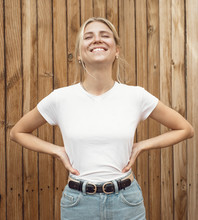 Young Pretty Girl Is Standing On A Light Wooden Wall Background. Woman Is Wearing A White Empty T-shirt Without Logo Which Makes It Being Perfectly Suitable For Mock-up.