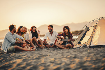 Happy friends sitting on the beach singing and playing guitar during the sunset