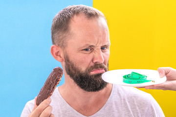 Diet and the problem of choice. Funny beard man doubts to eat an eclair seeing measuring tape