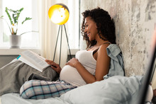 Cheerful pregnant woman reading a book in her bed.