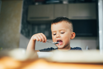 Wall Mural - Cute boy in the kitchen eating yogurt from a white container for yogurt, there is a place for advertising