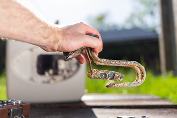 A man holding a water heater element damaged from corrosion. In the background the boiler with instrumets and a view of the lawn. Close up